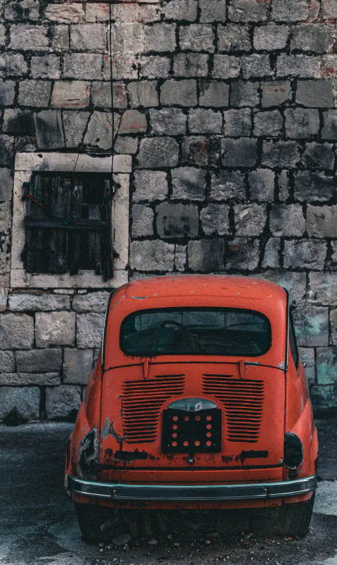 An old orange car parked against a weathered stone wall